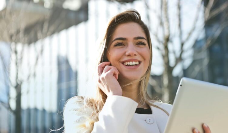 Confident businesswoman using her tablet and phone, smiling outdoors in sunlight.